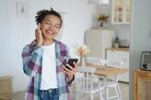 Happy mixed race girl in earphone holds smartphone using modern mobile apps, listening music at home photo