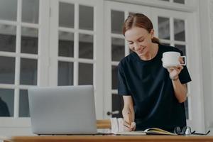 Female writing notes in notebook standing at desk, holding coffee mug, planning shedule in workplace photo