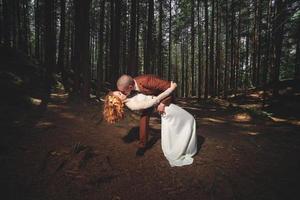 feliz pareja de recién casados con estilo en el bosque verde el día de verano. la novia con vestido blanco largo y el novio con traje rojo se abrazan. día de la boda. foto