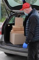 joven repartidor con máscara protectora, gorra roja y guantes cerca del camión con cajas y paquetes, al aire libre. coronavirus de servicio. las compras en línea. Bosquejo. foto