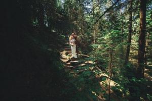 feliz pareja de recién casados con estilo en el bosque verde el día de verano. la novia con vestido blanco largo y el novio con traje rojo se abrazan. día de la boda. foto