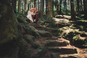 Happy stylish couple newlyweds in the green forest on summer day. bride in long white dress and groom in red suit are hugging. wedding day. photo