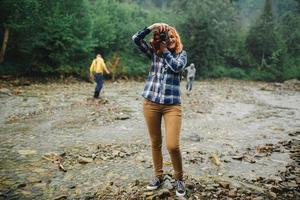 Young redhead woman with vintage camera takes photo near the river in a mountains