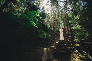 Happy stylish couple newlyweds in the green forest on summer day. bride in long white dress and groom in red suit are hugging. wedding day. photo