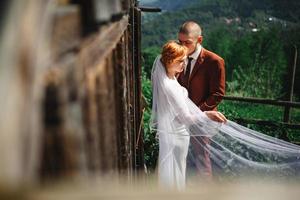 pareja de novios en las montañas. el novio y la novia se abrazan cerca de la casa rústica. una hermosa vista de las montañas detrás de la pareja foto