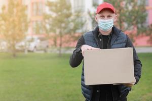 el repartidor con gorra roja, máscara médica facial sostiene una caja de cartón vacía al aire libre. coronavirus de servicio. las compras en línea. Bosquejo. foto