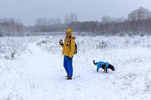 Young woman in yellow jacket with cup of coffee walking mixed breed Bedlington Whippet dog in blue warm suit on winter snowy day photo
