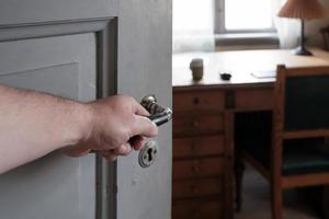 hhand sostiene la manija de una puerta de madera abierta, a través de la cual se puede ver una oficina, una mesa antigua, una silla, una lámpara y una ventana. foto