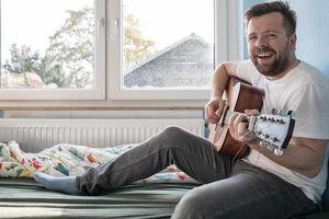 Man plays the acoustic guitar and sings while sitting on the bed. photo