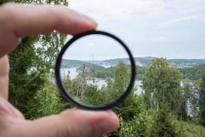 Photographer hand holds a polarizing filter, against the backdrop of trees, sky and sea. photo