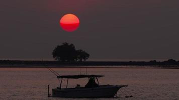 toneel- instelling zon visie, groot schijf gezien Bij horizon, reflectie streep twinkelen Aan water. donker silhouet van boot Aan voorgrond, wolken Bij horizon lijn video