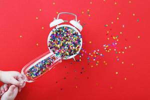 hands hold champagne glass with sequins pour on the alarm clock on a red background. christmas, celebration concept. photo