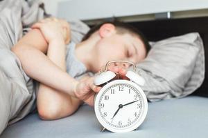 boy lying on the bed and stopping alarm clock in morning. childs hand reaching for the alarm clock to turn it off. photo