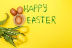 inscription happy easter from grass with bunny eggs, flowers tulips and nest on yellow background photo