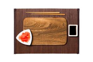 Empty rectangular brown wooden plate with chopsticks for sushi, ginger and soy sauce on dark bamboo mat background. Top view with copy space for you design photo