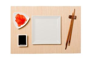 Empty white square plate with chopsticks for sushi and soy sauce, ginger on brown sushi mat background. Top view with copy space for you design photo