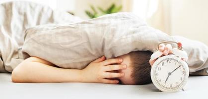 boy lying on the bed and stopping alarm clock in morning. childs hand reaching for the alarm clock to turn it off. photo