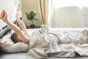 little boy lying and stretching in bed at home in the morning with alarm clock photo