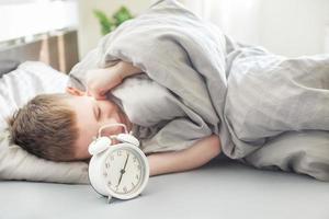 el niño se acuesta en la cama por la mañana y se cubre los oídos para no escuchar el despertador. niño no quiere despertar foto