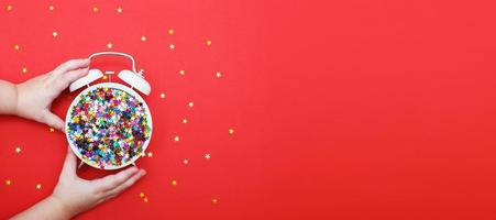 child's hands hold alarm clock with scattered sequins on a red background. christmas, celebration concept. banner photo