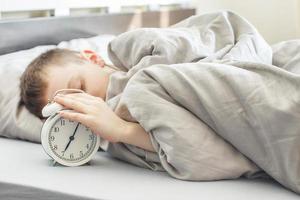 boy lying on the bed and stopping alarm clock in morning. childs hand reaching for the alarm clock to turn it off. photo