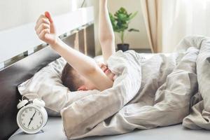 niño pequeño acostado y estirándose en la cama en casa por la mañana con despertador foto