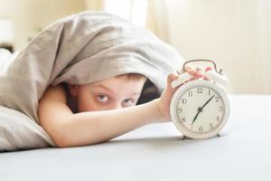 boy lying on the bed and stopping alarm clock in morning. childs hand reaching for the alarm clock to turn it off. photo
