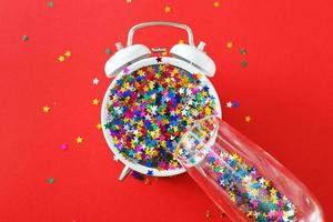 A glass with multicolored sequins, alarm clock and star shaped confettie on the red background. Party, new year, concept photo