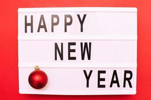 The inscription happy new year on a light box with Christmas toy ball on a red background. photo