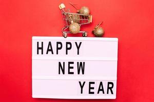 The inscription happy new year with shopping trolley with shiny gold christmas toys balls on a red background. photo