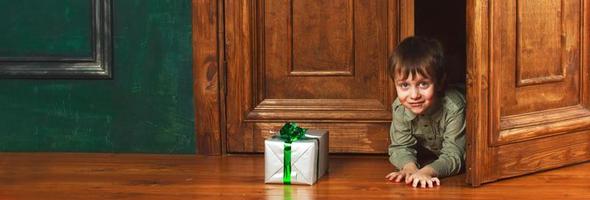 child boy peeks out of the door with a christmas gift box. banner photo