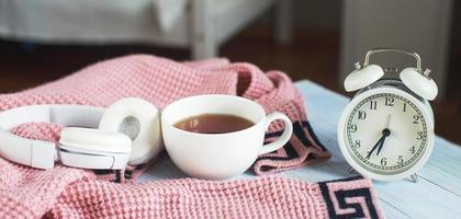 una taza de té caliente, suéter de punto, despertador y auriculares en el dormitorio. concepto de mañana de invierno. bandera foto