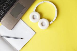 Top view open notepad, headphones pen and laptop on yellow background. workplace photo