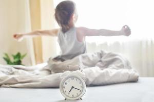 little boy sitting and stretching in bed at home in the morning on a window background with alarm clock photo