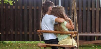 children sitting on a swing in the garden. older brother hugging little sister photo