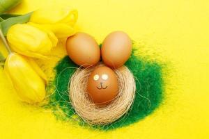 happy easter bunny eggs in nest with flowers tulips on the decorative grass on yellow background. copy space for text photo