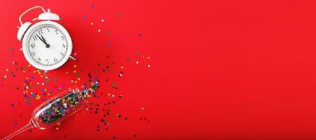 A glass with multicolored sequins, alarm clock and star shaped confettie on the red background. Banner photo