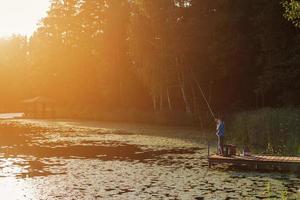 Little boy standing and fishing on a wooden dock at the sunset photo