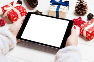 Top view of female hands holding a tablet on wooden Christmas background made of gift boxes. New year holiday concept. Mockup photo