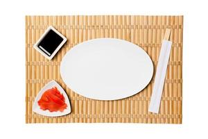 Empty oval white plate with chopsticks for sushi and soy sauce, ginger on yellow bamboo mat background. Top view with copy space for you design photo