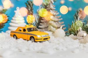 Christmas banner Background. Yellow toy car Taxi Cab model and winter decorations ornaments on blue background with snow and defocused garland lights. City traffic delivery taxi service concept. photo