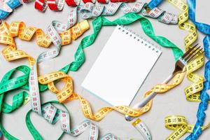 Composition of colorful measuring tapes, fork and notepad on cement background. Top view of diet control and empty space for your design photo