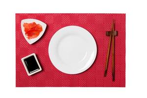 Empty round white plate with chopsticks for sushi and soy sauce, ginger on red mat sushi background. Top view with copy space for you design photo