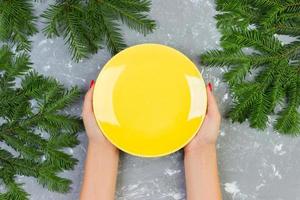 female hands holding an empty yellow plate on a gray background with Christmas branches. view from above for your design photo