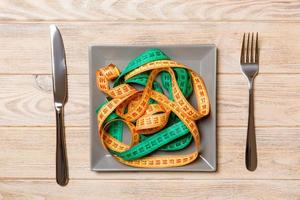 Top view of colorful measuring tapes on plate in the form of spaghetti with knife and fork on wooden background. Weight loss and diet concept photo