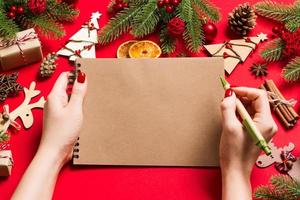 Top view of female hand making some notes in noteebok on red background. New Year decorations and toys. Christmas time concept photo