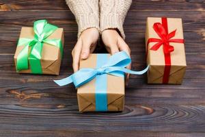 Woman hands give wrapped valentine or other holiday handmade present in paper with red, blue and green ribbon. Present box, decoration of gift on white wooden table, top view with copy space photo