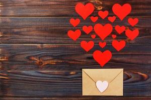 Valentine day love letter. Envelope from craft paper with red hearts heap spread on white wood background. Lover's holiday confession or proposal concept photo