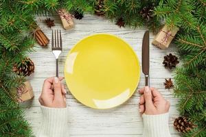 top view girl holds fork and knife in hand and is ready to eat. Empty plate round ceramic on wooden christmas background. holiday dinner dish concept with new year decor photo