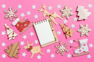 Top view of notebook, pink background decorated with festive toys and Christmas symbols reindeers and New Year trees. Holiday concept photo